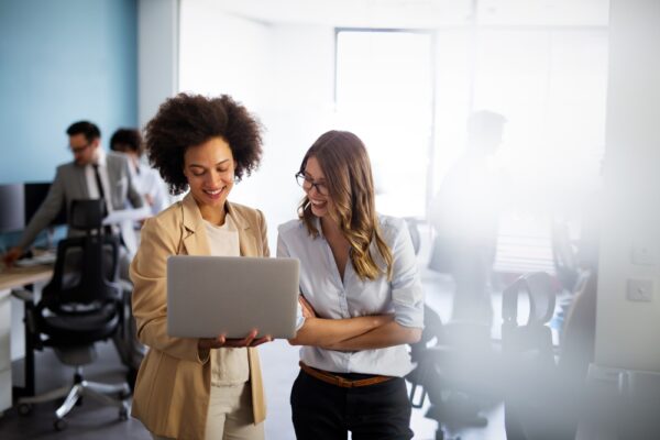 Two female colleagues smiling and looking at a laptop in a bright, modern office setting, representing teamwork and professional SEO strategy planning.