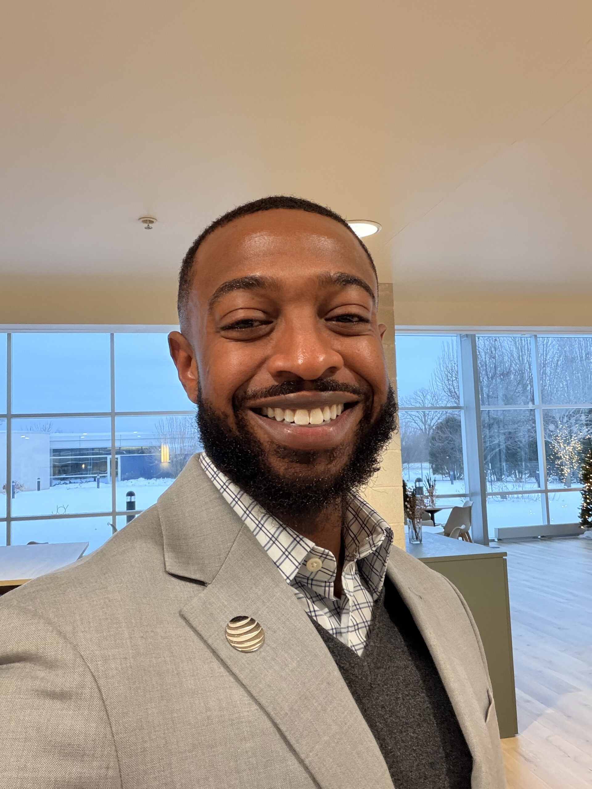 A smiling professional man in a grey blazer and button-down shirt standing in a modern, sunlit office space, representing approachable business leadership.