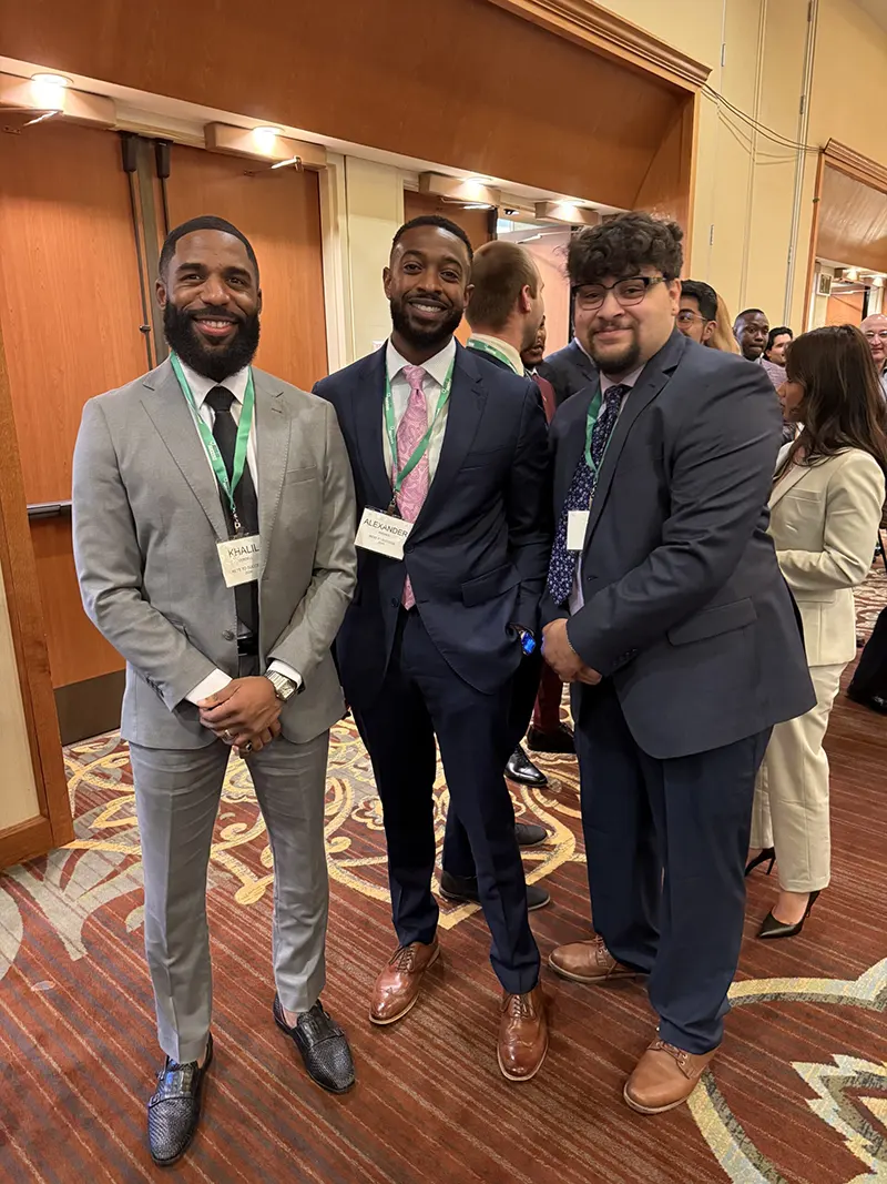 Three diverse professional men in business suits wearing event lanyards at a corporate networking conference, illustrating professional growth and team collaboration.