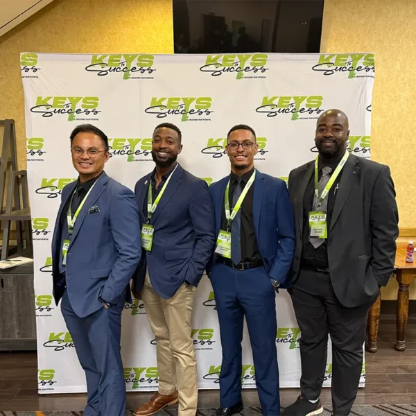 Four professional men in business suits standing in front of a Keys to Success backdrop at a corporate networking event.
